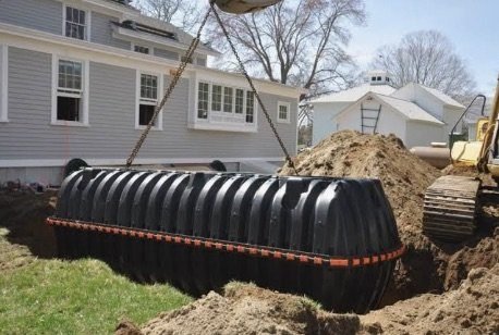 Septic tank being lowered by crane into an excavated trench beside a house